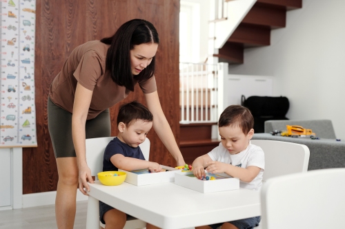 young woman watching two boy toddlers doing an activity at a small table
